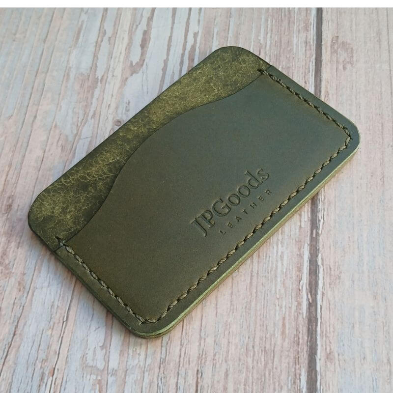 Vihreä nahkainen korttikotelo, jossa JPGOODS-logo, asetettuna vaalealle puulaudalle.